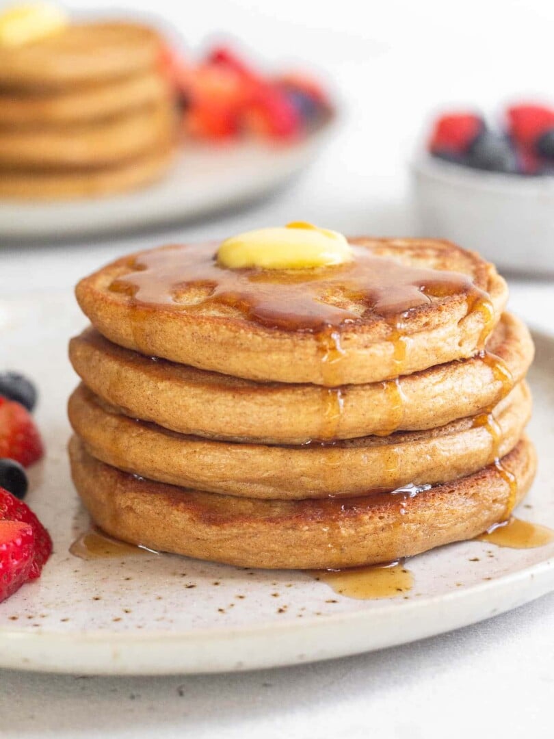 A plate of four cottage cheese pancakes topped with butter and syrup. There are some fresh berries on the plate. Behind the plate is a bowl of more berries and another stack of pancakes.