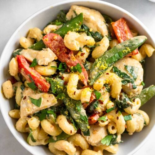 A bowl of tahini pasta with chicken and veggies. It is garnished with fresh herbs. Around the bowl is half a lemon, a ramekin of spices, and a pan.