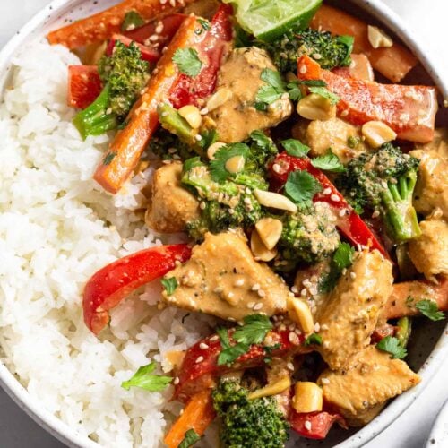 Peanut butter chicken in a bowl with white rice topped with cilantro, peanuts, sesame seeds and a lime wedge. Behind the bowl is a kitchen towel and a small bowl of peanuts.
