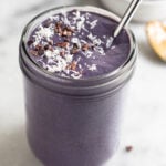 Healthy smoothie in a jar topped with coconut shreds and cacao nibs with a straw. Behind it is a bowl of frozen blueberries and a spoon with peanut butter on it.