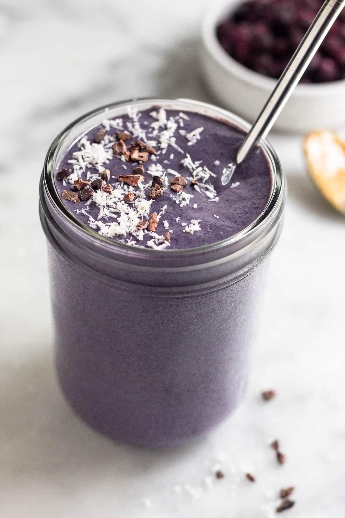 Healthy smoothie in a jar topped with coconut shreds and cacao nibs with a straw. Behind it is a bowl of frozen blueberries and a spoon with peanut butter on it.