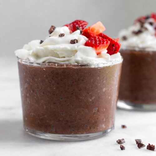 Chocolate chia pudding in a small glass topped with whipped cream, strawberries, and cacao nibs. Behind it is a similar glass and around them is cacao nibs.