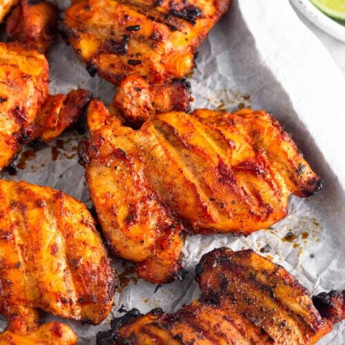 Grilled honey chipotle chicken thighs on a baking sheet lined with parchment paper. Next to it is a plate of lime wedges.