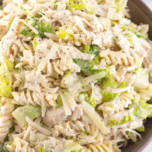 A close up of a bowl of chicken caesar pasta salad. It is garnished with black pepper. Behind the bowl is a plate of grated parmesan cheese.
