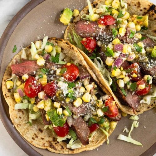 A plate of two flank steak tacos stuffed with cabbage, grilled steak, corn, tomatoes, avocado, salsa, cheese, and cilantro. Around the plate is a plate of lime wedges, a bowl of salsa, and a plate of tortillas.