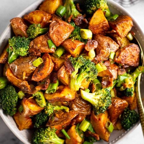 Large bowl filled with roasted potato salad with bacon and broccoli with a spoon in it. Next to the bowl is a striped towel.