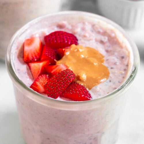 A jar of strawberry overnight oats topped with fresh diced strawberries and peanut butter. Around it is another jar of overnight oats and a small dish of strawberries.