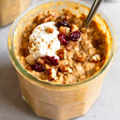 Two jars of pumpkin overnight oats, with one being behind the other. They are topped with greek yogurt, chopped pecans, dried cranberries, and cinnamon. A spoon is in the jar in the front. A ramekin of pecans is off to the side of the one in the back.