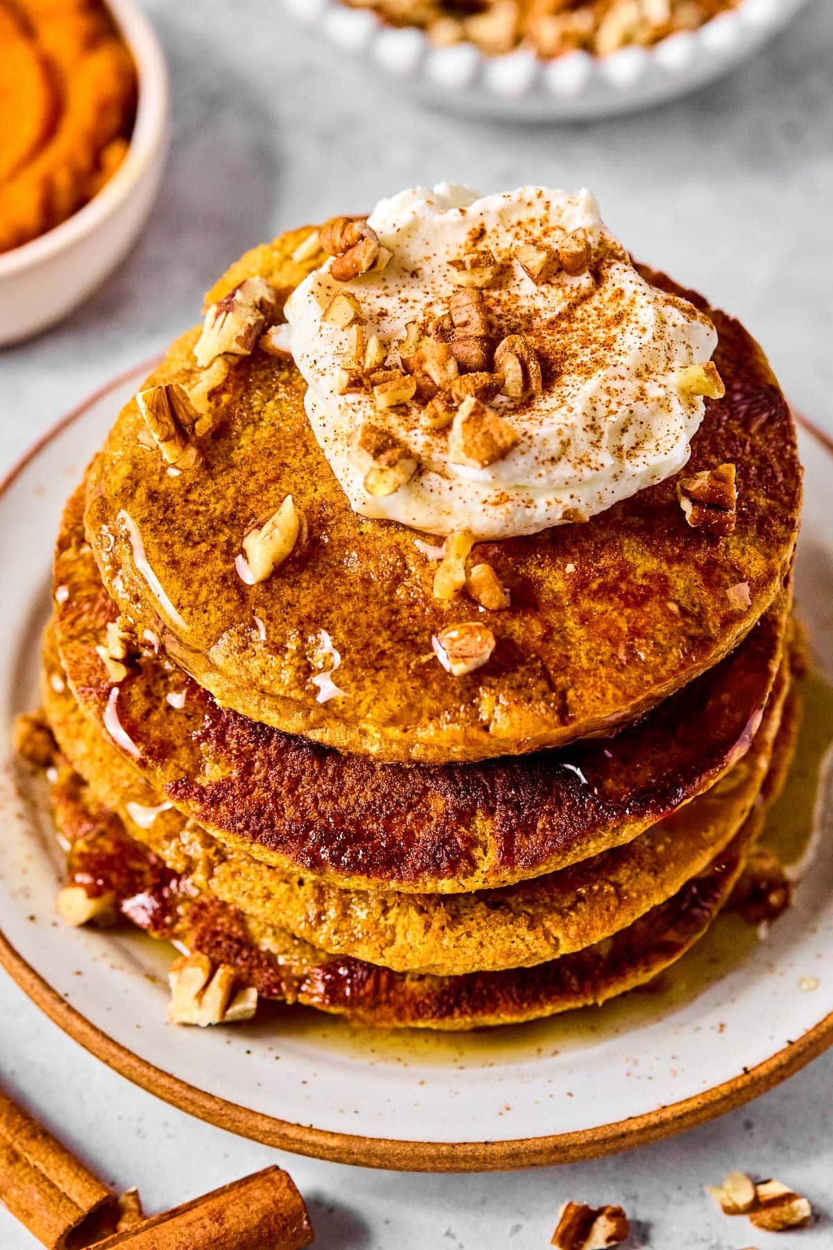 A stack of pumpkin pancakes topped with greek yogurt, pecan pieces, cinnamon, and maple syrup on a plate. Around the plate are some cinnamon sticks, pecan pieces, a bowl of pumpkin puree, and a bowl of pecans.