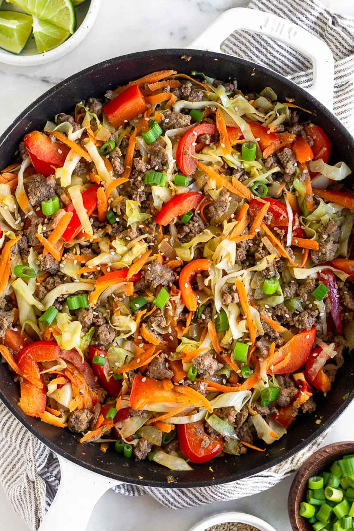 Healthy one pan meal with ground beef, shredded cabbage, bell peppers, and carrots in a large cast iron skillet. It is garnished with green onions. Next to the pan is a bowl of green onions and a bowl of lime wedges.