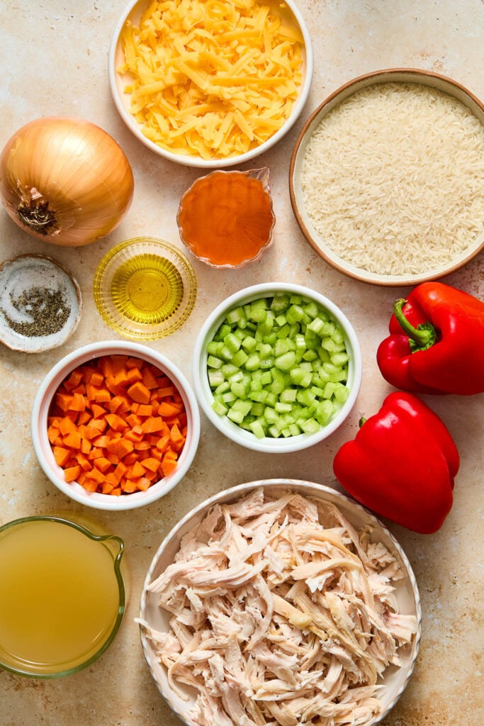 A bowl of shredded orange cheese, a bowl of uncooked white rice, two red bell peppers, a bowl of shredded chicken, a measuring cup of broth, a bowl of diced red bell pepper, a bowl of diced celery, a cup of buffalo sauce, a small bowl of olive oil, a small bowl of salt and pepper, and a whole yellow onion.