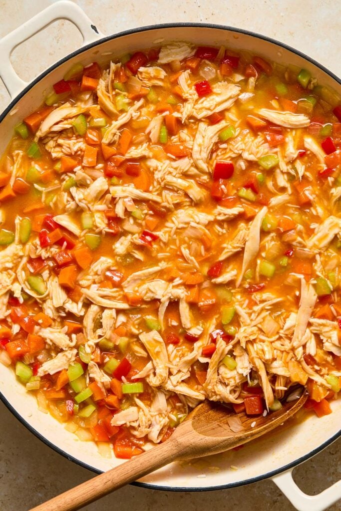 Large white enameled skillet filled with a mixture of saut&eacute;ed veggies, shredded chicken, uncooked rice, buffalo sauce, and broth.