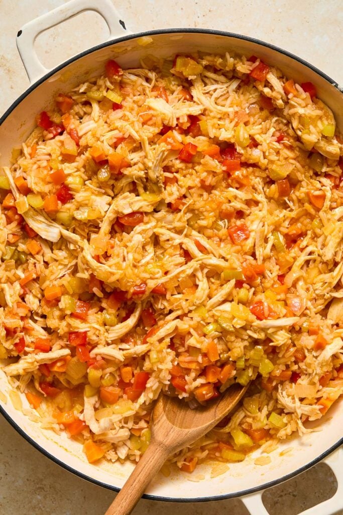 Large white enameled skillet filled with a cooked mixture of shredded chicken, white rice, and veggies. A wooden spoon is also in the skillet.