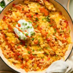 Buffalo chicken casserole in a large white enameled skillet topped with melted cheese, a dollop of greek yogurt, sliced green onions, and hot sauce. A kitchen towel is tied around the handle. Around the pan is a plate with two white forks, a small dish of sliced green onions, a bowl of shredded cheese, and another plate.