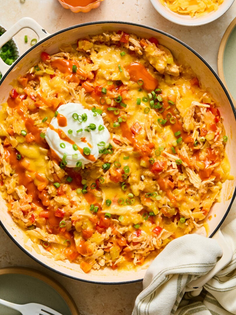 Buffalo chicken casserole in a large white enameled skillet topped with melted cheese, a dollop of greek yogurt, sliced green onions, and hot sauce. A kitchen towel is tied around the handle. Around the pan is a plate with two white forks, a small dish of sliced green onions, a bowl of shredded cheese, and another plate.