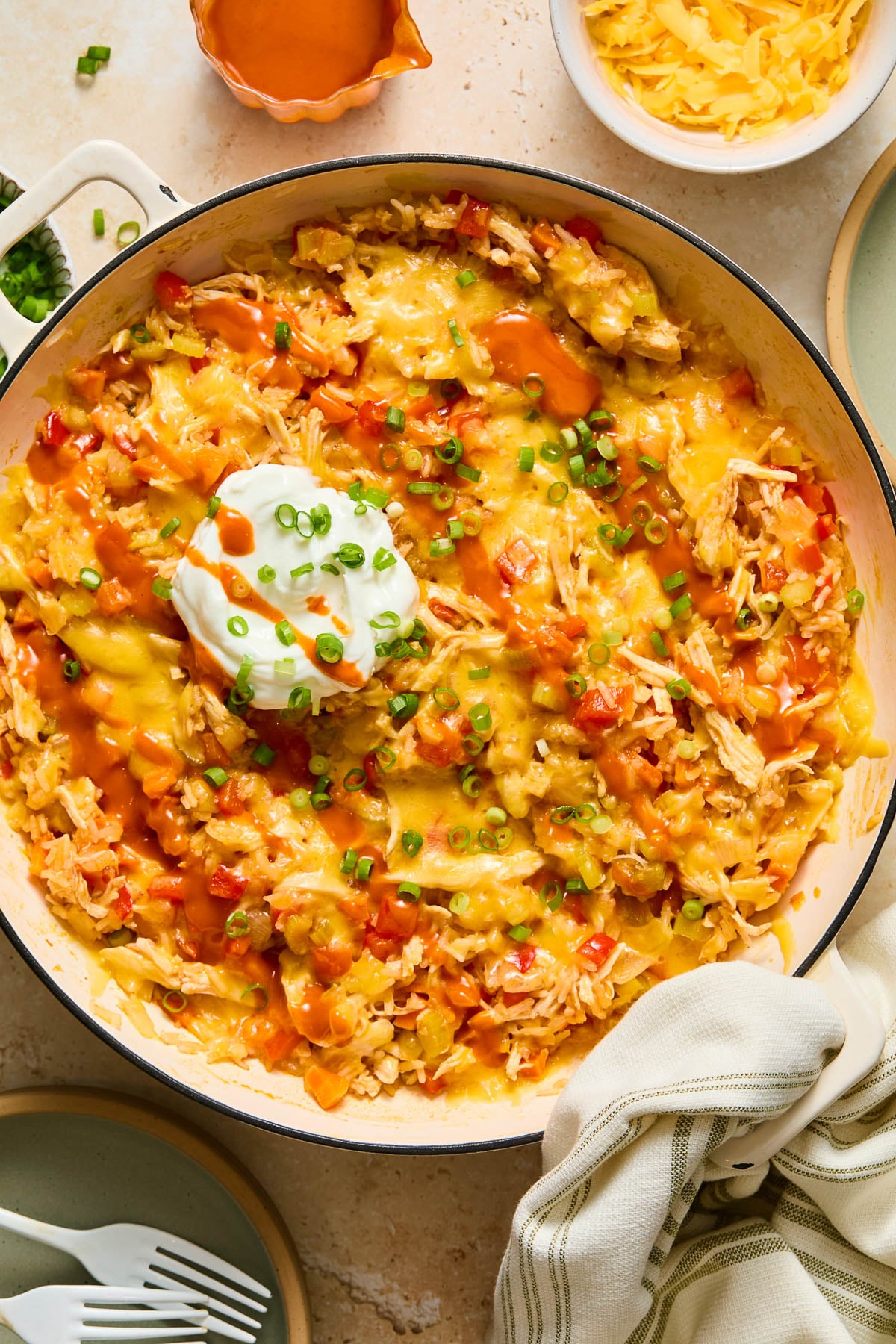 Buffalo chicken casserole in a large white enameled skillet topped with melted cheese, a dollop of greek yogurt, sliced green onions, and hot sauce. A kitchen towel is tied around the handle. Around the pan is a plate with two white forks, a small dish of sliced green onions, a bowl of shredded cheese, and another plate.