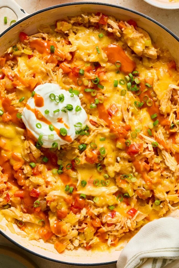 Cheesy buffalo chicken and rice casserole in a large white enameled skillet. It is topped with a big dollop of greek yogurt, sliced green onions, and drizzled with hot sauce.