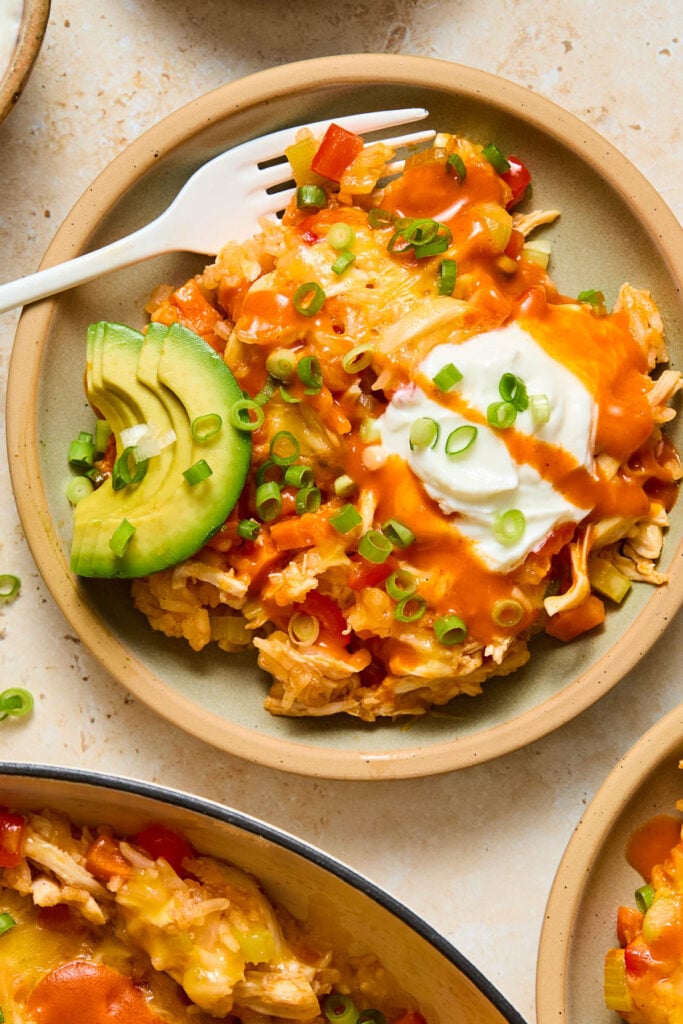 A plate of cheesy buffalo chicken casserole topped with greek yogurt, sliced green onions, hot sauce, and sliced avocado. A fork is resting on the plate too. Around the plate is another plate of casserole and a pan filled with the remaining casserole.