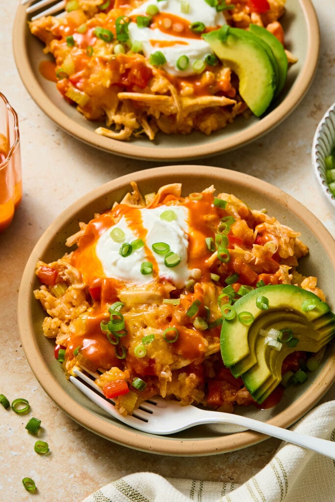 Two plates of healthy buffalo chicken rice casserole with melted cheese. They are topped with greek yogurt, hot sauce, green onions, and sliced avocado. A white fork is resting on the plate in front. Around the plates is a kitchen towel, some sliced green onions, a cup of buffalo sauce, and a bowl of more sliced green onions.