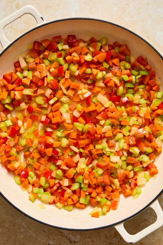 Large white enameled skillet filled with diced saut&eacute;ed onions, carrots, celery, and red bell peppers.
