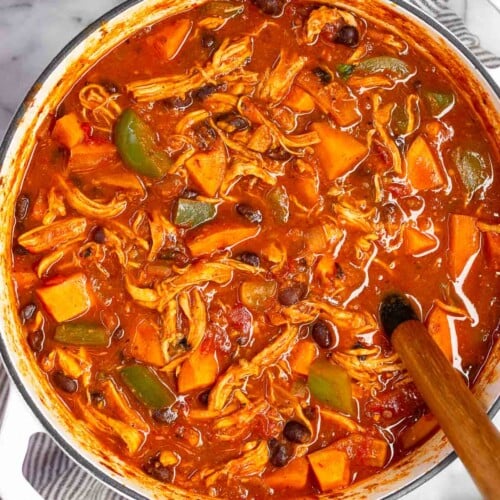 Chicken sweet potato soup in a large white dutch oven with a spoon in it. The pot is sitting on a striped towel with half an avocado, a bowl of cilantro, and a plate of lime wedges around it.