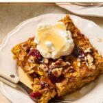 A piece of pumpkin baked oatmeal with cranberries and pecans on a white plate. It is topped with greek yogurt and a drizzle of maple syrup. A little piece is taken off the corner and is sitting on a fork on the plate. Around the plate is a pink napkin, a dish of dried cranberries, and another plate.