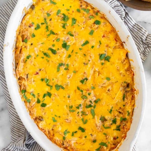White baking dish with cheesy chicken dip garnished with cilantro. The dish is on a striped kitchen towel with a bowl of tortilla chips next to it.