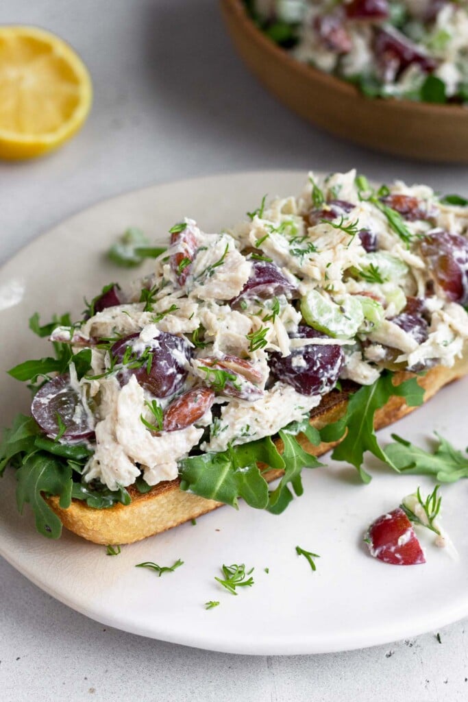 High protein chicken salad with grapes, celery, and almonds on top of some arugula on a piece of toast on a plate. Behind it is half a lemon and a bowl of more chicken salad.