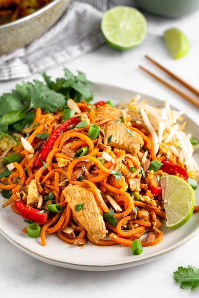 Plate with sweet potato noodle chicken pad thai garnished with a lime wedge, cilantro, and bean sprouts. Around it is more lime wedges, cilantro, and chopsticks.