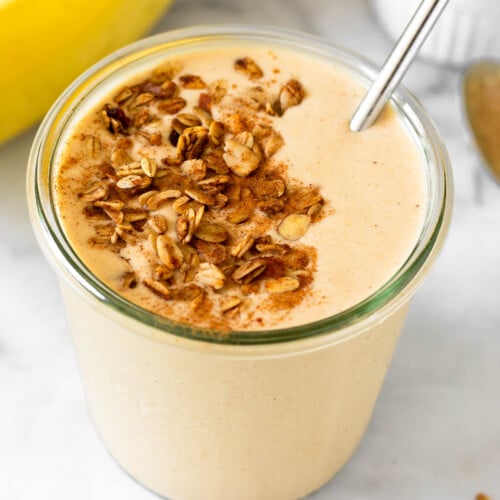 Peanut butter banana protein smoothie in a glass jar topped with granola and cinnamon with a straw in it. Around the jar are some bananas and a bowl of granola.