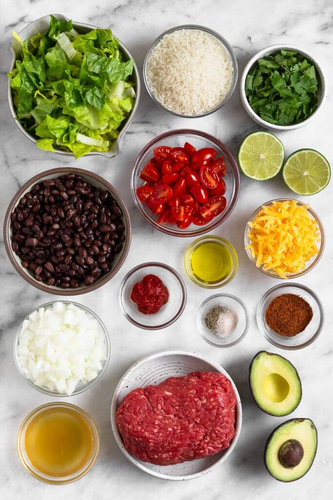 White marble counter with a bowl of uncooked white rice, a bowl of cilantro, a lime cut in half, a bowl of shredded cheddar cheese, a bowl of diced tomatoes, a bowl of olive oil, a bowl of salt and pepper, a bowl of spices, an avocado that has been cut in half, a bowl of raw ground beef, a bowl of broth, a bowl of diced white onion, a bowl of tomato paste, a bowl of black beans, and a bowl of chopped romaine.