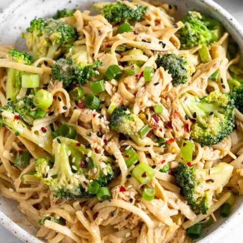 Sesame noodles with chicken and broccoli garnished with green onion, sesame seeds, and red pepper flakes in a white bowl.