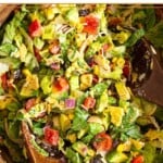 Southwest chicken salad with lettuce, shredded chicken, tomatoes, peppers, beans, corn, avocado, crushed tortilla chips, and red onion tossed with a salsa dressing in a large wooden salad bowl with two wooden spoons in it.