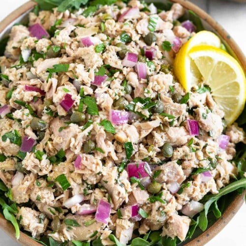 Italian tuna salad with capers in a bowl with arugula underneath it. It is garnished with 2 lemon wedges. Around the bowl is a half of lemon and 2 slices of bread.