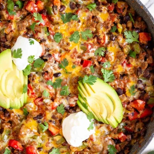 Taco skillet topped with melted cheese, sliced avocado, sour cream, and cilantro in a large sauté pan. Next to the skillet is a bowl of cilantro, two lime wedges, and a bowl of tortilla chips.