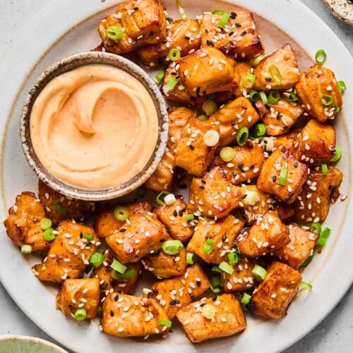 A plate of air fry salmon bites topped with diced green onions and sesame seeds. Also on the plate is a bowl of sriracha mayo for dipping. Around the plate is a bowl or more diced green onions and two different bowls of sesame seeds.