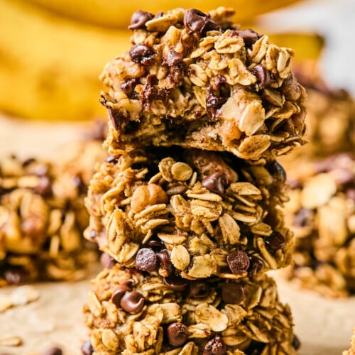 Three banana oatmeal cookies stacked on top of each other. The top one has a bite taken out of it. Around them are more cookies and in the far distance are some bananas.