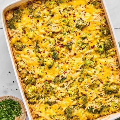 Healthy chicken broccoli rice casserole covered with melted cheese and topped with red pepper flakes and fresh parsley in a large baking dish. Next to the dish is a small bowl of red pepper flakes, a small bowl of parsley, and a bowl of shredded cheese.