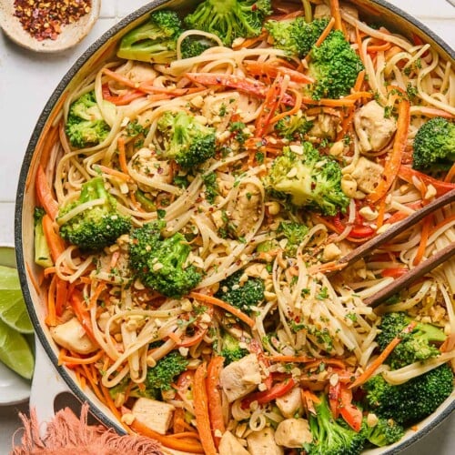 Peanut butter noodles with chicken and veggies in a large white skillet. It is garnished with cilantro and chopped peanuts and also in the pan is a pair of chopsticks. The handle of the pan is wrapped in a red towel and around the pan is a plate of lime wedges, a bowl of red pepper flakes, and a bowl of chopped peanuts.