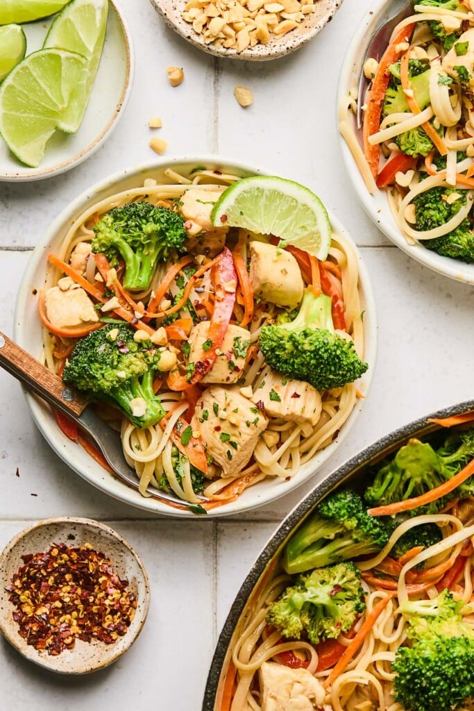 A plate of peanut butter noodles with chicken, broccoli, red bell peppers, and carrots garnished with cilantro, crushed peanuts, and red pepper flakes. The plate as a lime wedge on it and also a fork. Around the plate is a bowl of red pepper flakes, a plate of lime wedges, a bowl of crushed peanuts, another plate of peanut noodles, and a pan of peanut noodles.