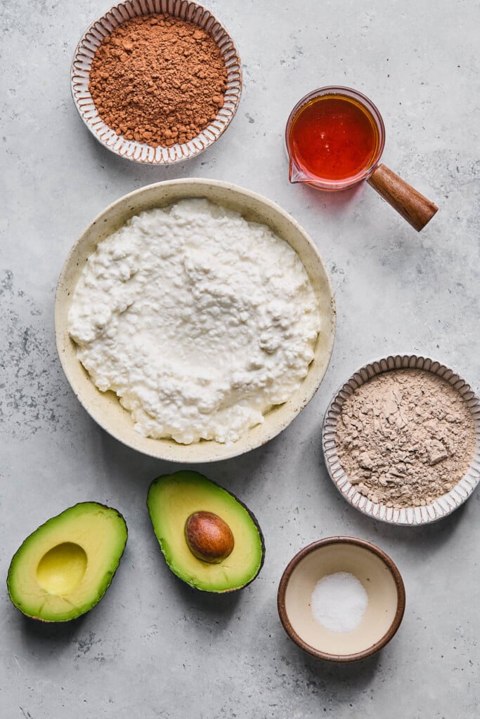 Grey concrete counter with a cup of maple syrup, a bowl of cottage cheese, a bowl of chocolate collagen, a small bowl of salt, an avocado that has been cut in half, and a bowl of cocoa powder.