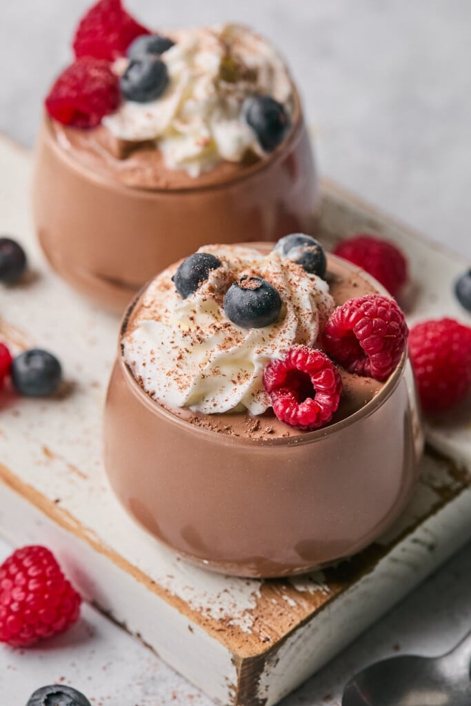 Two glass cups of chocolate protein pudding topped with whipped cream, fresh berries, and cinnamon. The cups are sitting on a white wooden board and around the cups is more fresh berries.