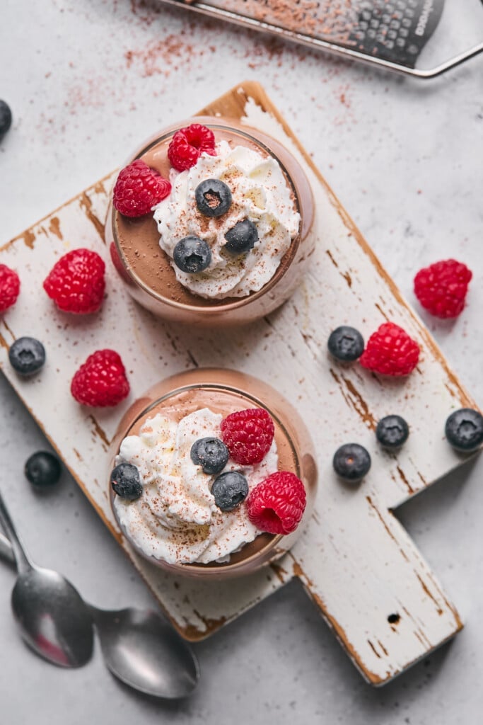 Overhead shot of two cups of cottage cheese chocolate pudding topped with whipped cream, berries, and cinnamon on a white cutting board. Around the cups are some more berries and two spoons.