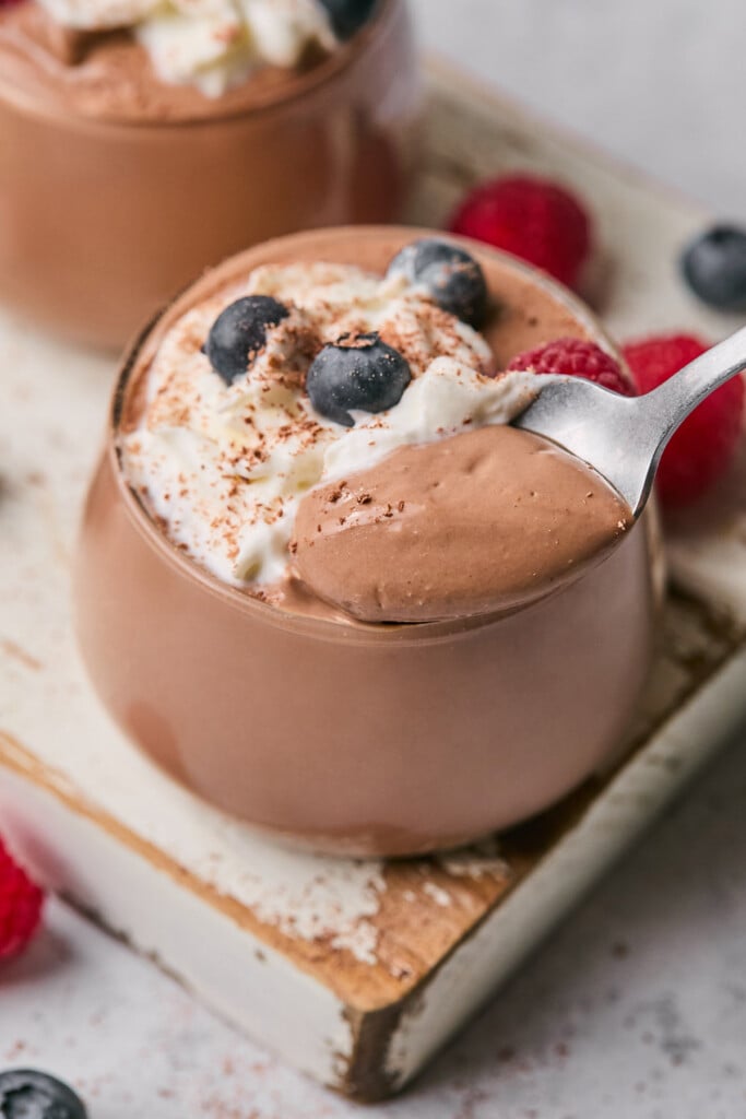 Cottage cheese pudding in a cup topped with whipped cream, berries, and cinnamon. A spoon with a scoop of pudding is being held overtop the jar. Around the jar is another cup of pudding and some fresh fruit.