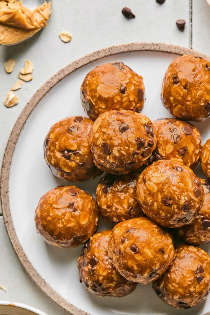 Peanut butter protein balls with chocolate chips stacked on top of each other on a white plate. Around the plate is a spoonful of peanut butter, some rolled oats, and a few mini chocolate chips.