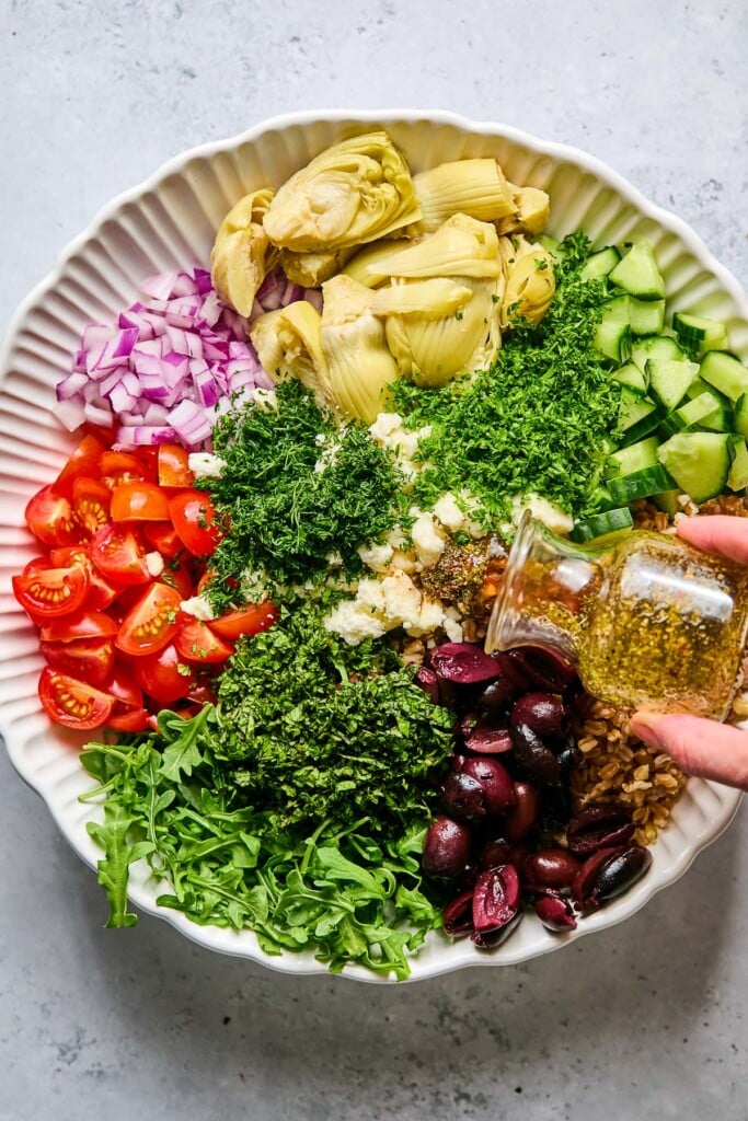 Large bowl with arugula, diced tomatoes, diced red onion, quartered artichoke hearts, diced cucumber, cooked farro, Kalamata olives, crumbled feta, and fresh herbs in it before it is mixed together. Dressing is being poured overtop of it.
