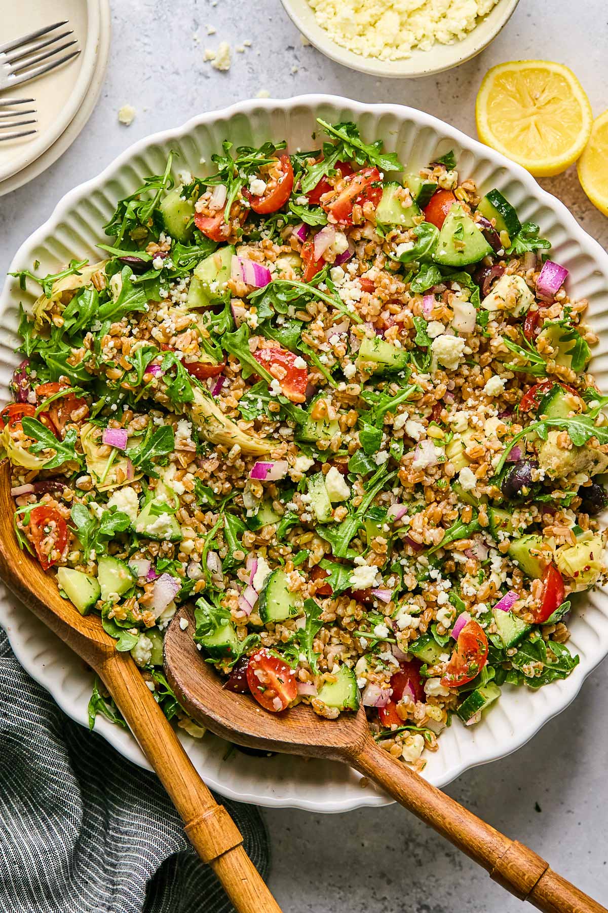 Large bowl filled with Mediterranean farro with two wooden salad tongs in the bowl. Around the bowl is a lemon cut in half, a striped linen towel, and a bowl with feta in it.