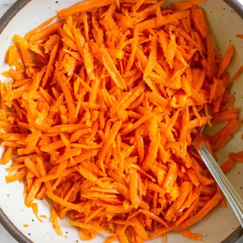 Raw carrot salad in a white bowl with a fork in it. Next to the bowl is a striped linen towel.
