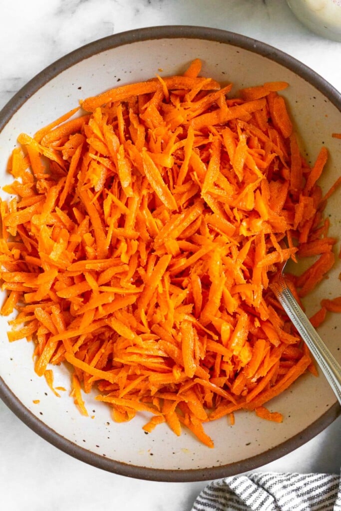 Raw carrot salad in a white bowl with a fork in it. Next to the bowl is a striped linen towel.