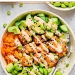 Salmon rice bowl with cubed salmon, white rice, edamame, diced avocado, shredded carrots, and diced cucumber with a fork in the bowl. Everything is drizzled with sriracha mayo and garnished with sesame seeds and green onions. Around the bowl is a bowl of diced cucumber and two bowls of edamame.